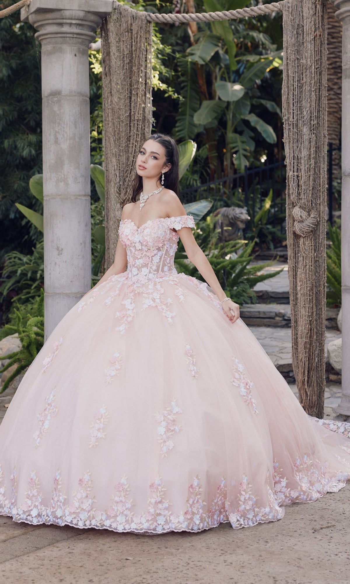 A woman stands outdoors in the Quinceanera Dress By Juliet JT1460J, a blush pink, off-the-shoulder ball gown with floral embellishments and a full skirt, surrounded by greenery and stone pillars.